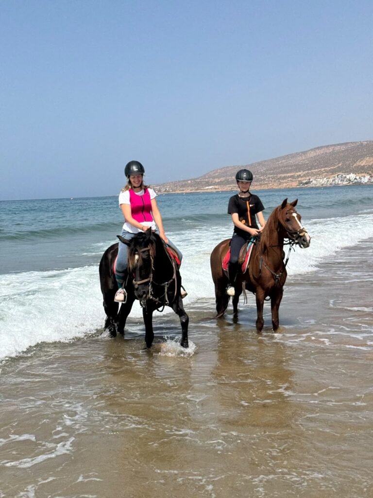 Horse riding in Agadir — two riders taking horses through the shallows on the beach below the headland